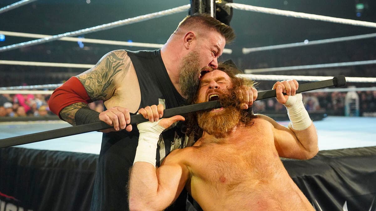 Kevin Owens pressing a bar into Sami Zayn's mouth while biting the top of his head