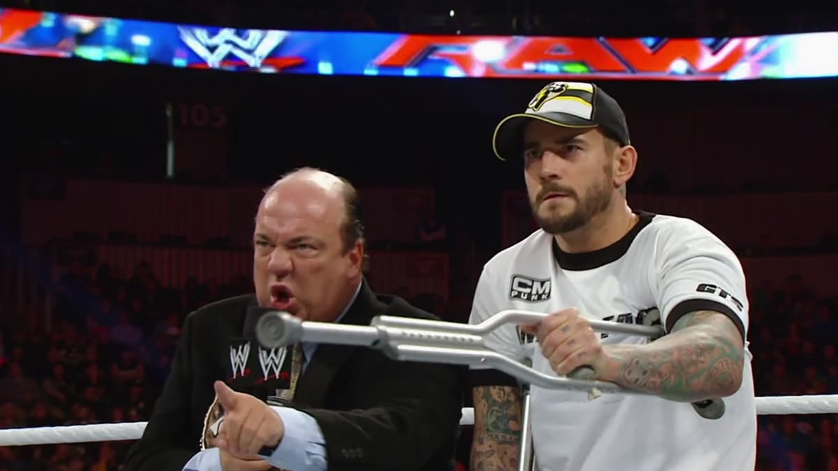 Paul Heyman angrily shouting into a microphone as CM Punk points on crutches 