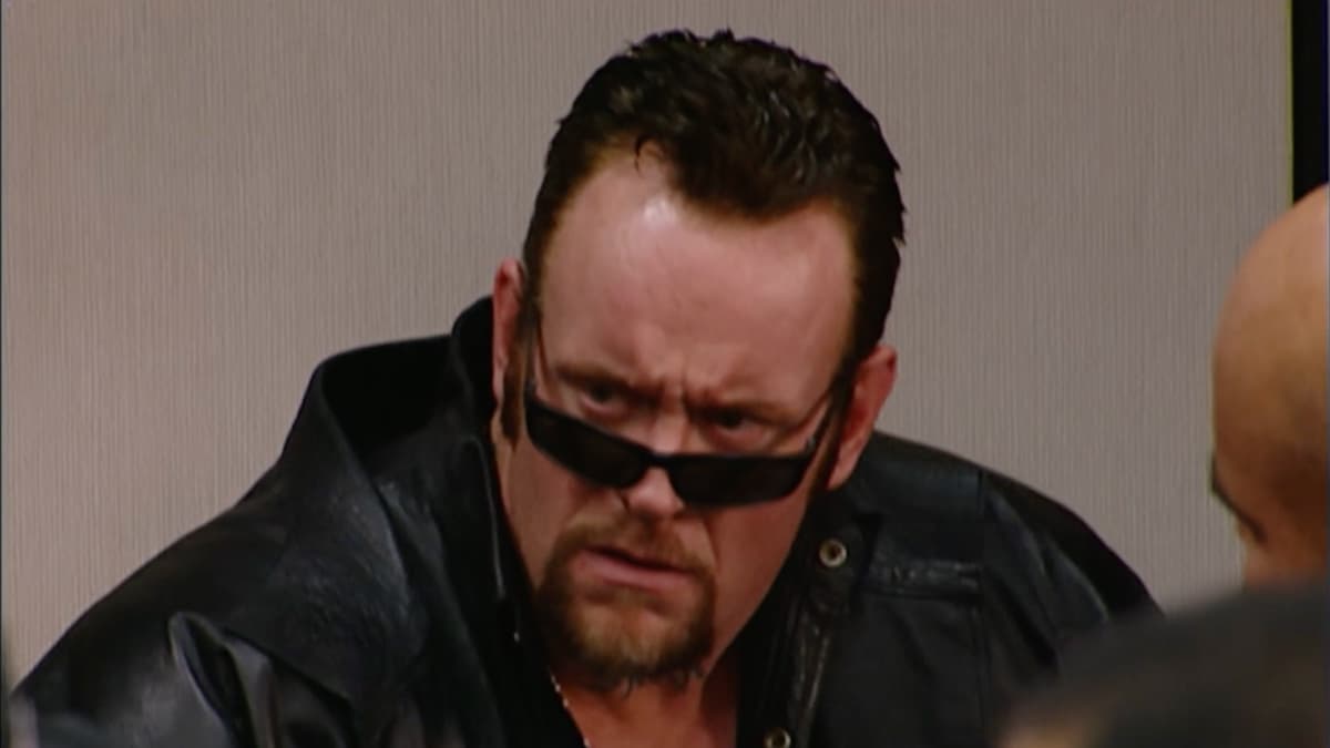 The Undertaker, wearing a leather jacket and with his sunglasses slightly lowered looking disgusted