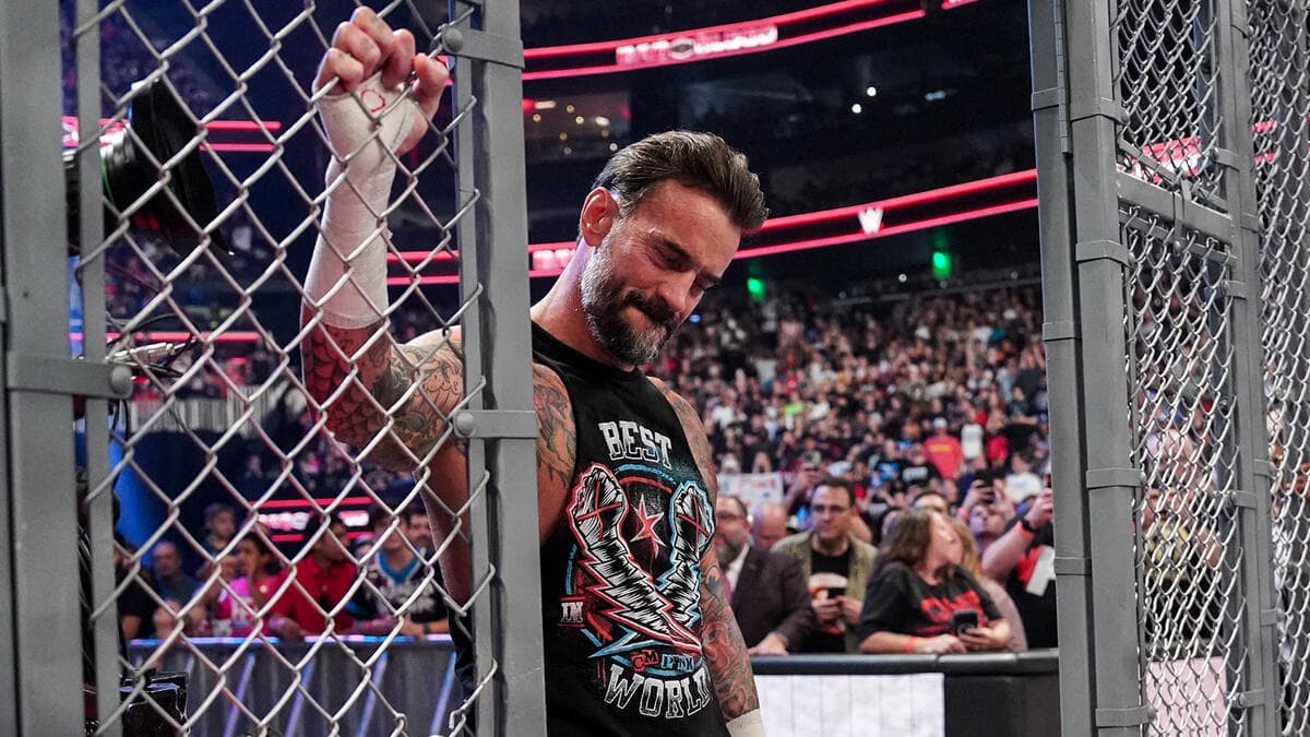 CM Punk with his head down wearing a black vest and holding the side of metal grating that makes part of WWE Hell in a Cell
